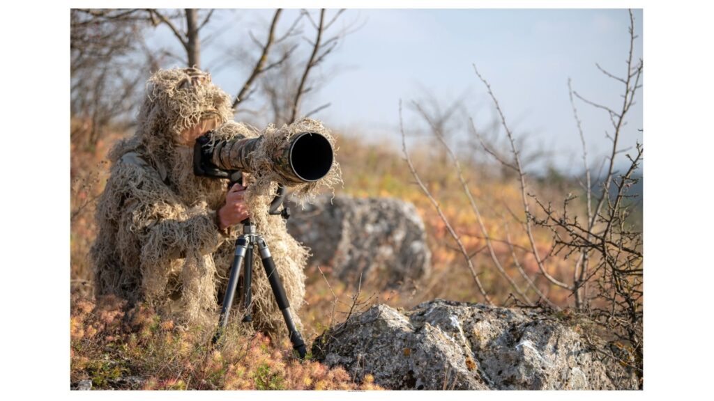 Wildlife photographer in full ghillie suit using tripod and telephoto lens to photograph wildlife in natural terrain